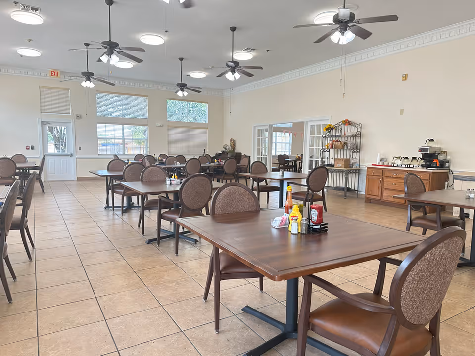 A spacious dining room with multiple wooden tables and cushioned chairs arranged neatly. The room has large windows allowing natural light, ceiling fans with lights, and a tiled floor. There is a sideboard with coffee machines and condiments on the tables.