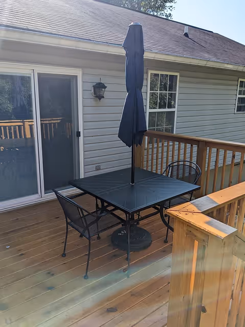 Wooden outdoor deck with a black patio table, umbrella, and two metal chairs next to a house and sliding glass door.