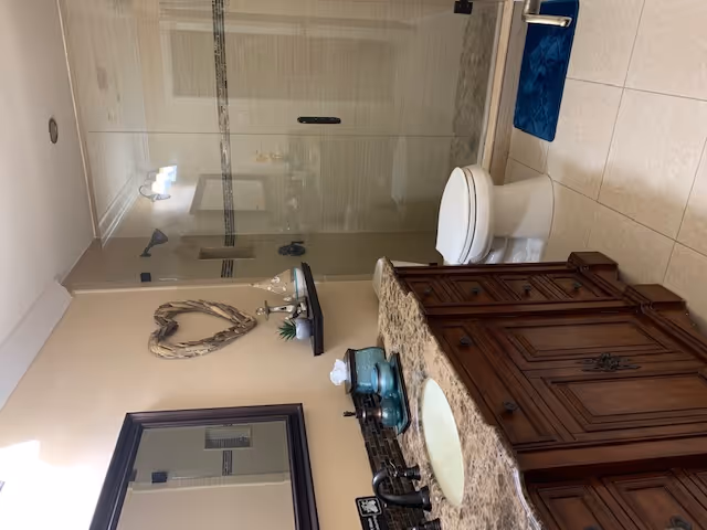 A bathroom with a wooden vanity topped with a granite countertop, a sink, and various decorative items including a small plant and candles. There is a large mirror above the vanity and a heart-shaped wall decoration. The bathroom also features a toilet and a glass-enclosed shower with tiled walls and a blue bath mat on the floor.