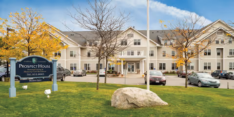 Front exterior of Prospect House Assisted Living and Memory Care, a three-story beige building with a lawn, sign, parked cars, and trees under a blue sky.