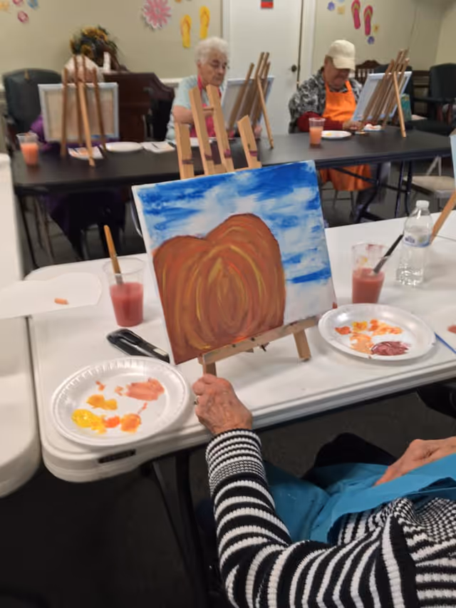 A group of elderly individuals seated at tables in a room, engaged in a painting activity. One person in the foreground is painting on a canvas with brown and orange colors against a blue and white background. Paint palettes, brushes, and cups with paint are visible on the tables. The room has light-colored walls decorated with flower and flip-flop cutouts.