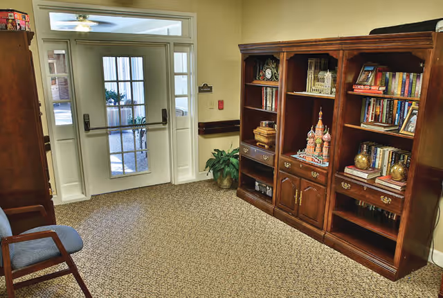 Carpeted entry area with glass double doors, wooden bookcases filled with books and decor, and a chair.