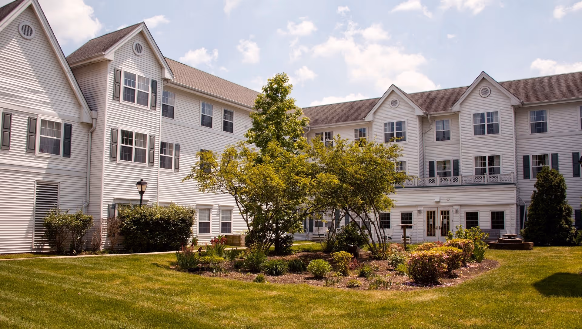 White three-story senior living building with multiple windows facing a landscaped grassy courtyard centered on a small tree.