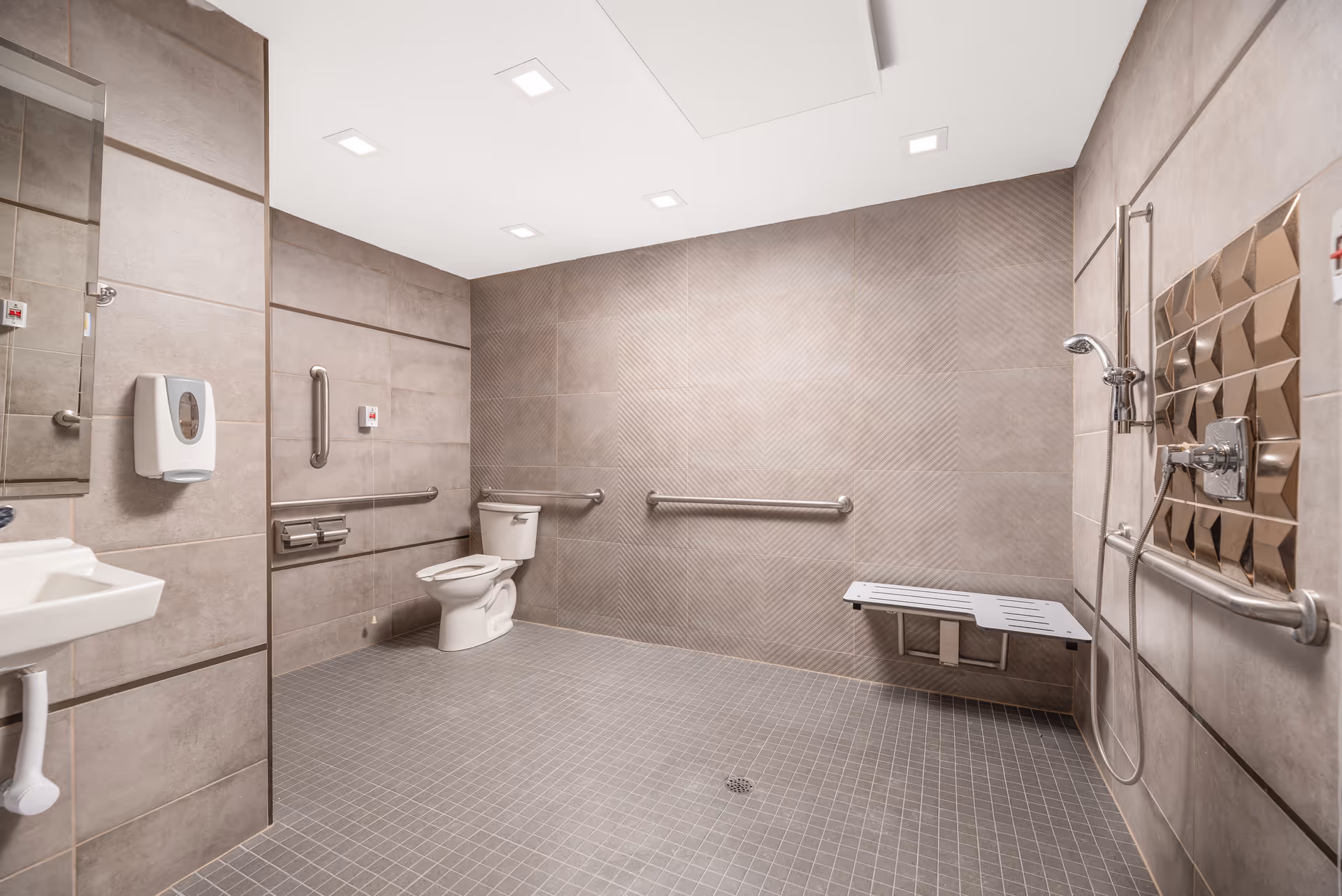 Spacious accessible bathroom with beige tiled walls and floor, featuring a toilet with grab bars, a wall-mounted sink, a soap dispenser, a shower area with a handheld showerhead, grab bars, and a fold-down shower bench.