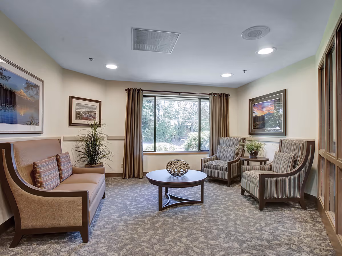 A cozy sitting area with a beige sofa and two striped armchairs arranged around a round wooden coffee table. The room has a large window with brown curtains, framed landscape pictures on the walls, and a patterned carpet. There are also two potted plants placed on side tables.