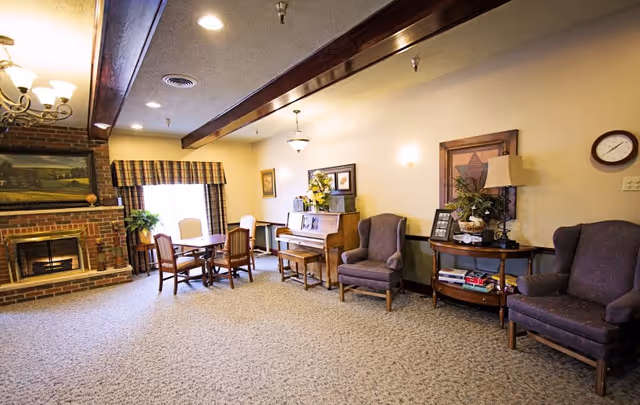 A cozy living room area in Elms Retirement Village featuring a brick fireplace with a painting above it, a wooden table with four chairs near a window with plaid curtains, a piano with a floral arrangement on top, two upholstered armchairs, and a wooden side table with a lamp, books, and framed pictures. The room has beige walls, carpeted floor, and exposed wooden ceiling beams.