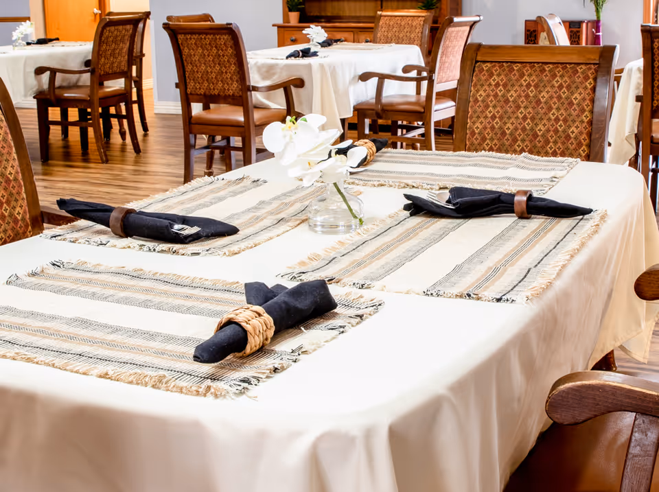 A dining area with tables covered in white tablecloths and striped placemats. Each place setting has a black cloth napkin with a napkin ring. Wooden chairs with patterned upholstery surround the tables. A small vase with white flowers is placed in the center of the table in the foreground. The floor is wooden, and additional tables and chairs are visible in the background.