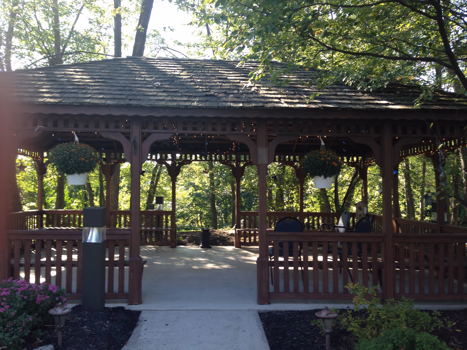 Wooden gazebo with hanging flower baskets and seating surrounded by trees.
