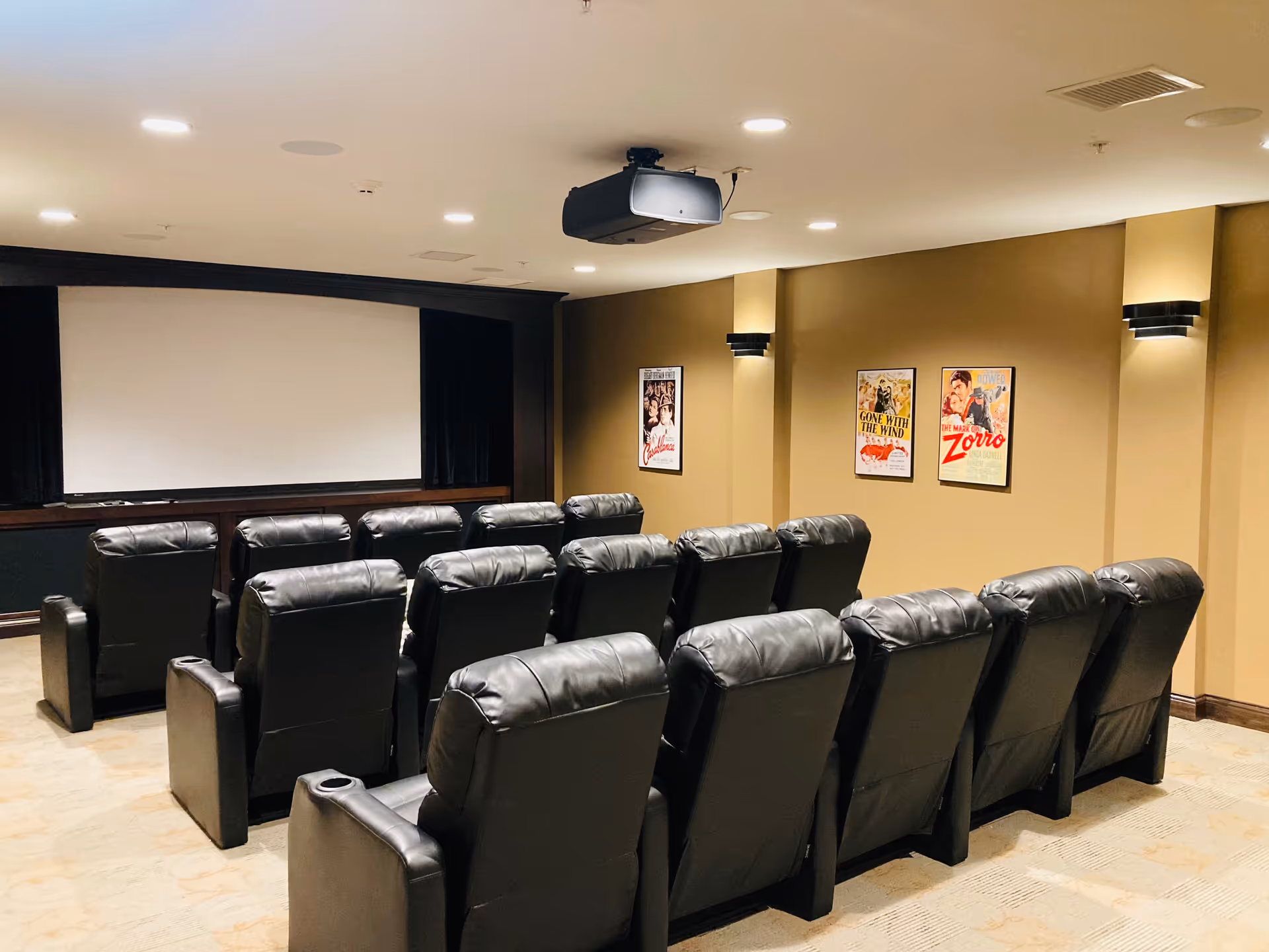 A small movie theater room with three rows of black leather recliner chairs facing a large blank projection screen. The walls are beige with three framed vintage movie posters and modern wall sconces providing soft lighting. A ceiling-mounted projector is positioned above the chairs.