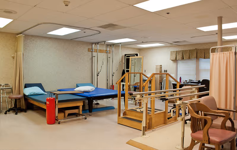 A physical therapy room with a blue therapy table, wooden parallel bars with stairs, a wooden chair with a pink cushion, a walker, and various therapy equipment. The room has beige walls, a tiled ceiling with fluorescent lights, and windows with valances.