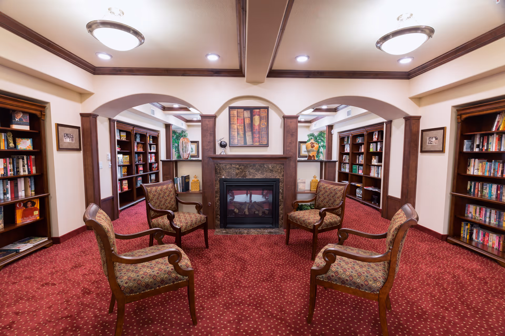 A cozy library room with four upholstered armchairs arranged around a central fireplace. The room features red carpet with a dotted pattern, wooden bookshelves filled with books along the walls, and decorative items on the shelves. The ceiling has recessed lighting and two circular light fixtures, with wooden trim accents throughout the space.