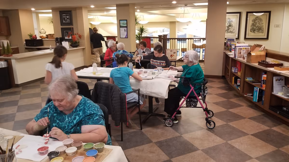 Older adults and a few children sit at tables in a communal indoor activity room painting crafts.
