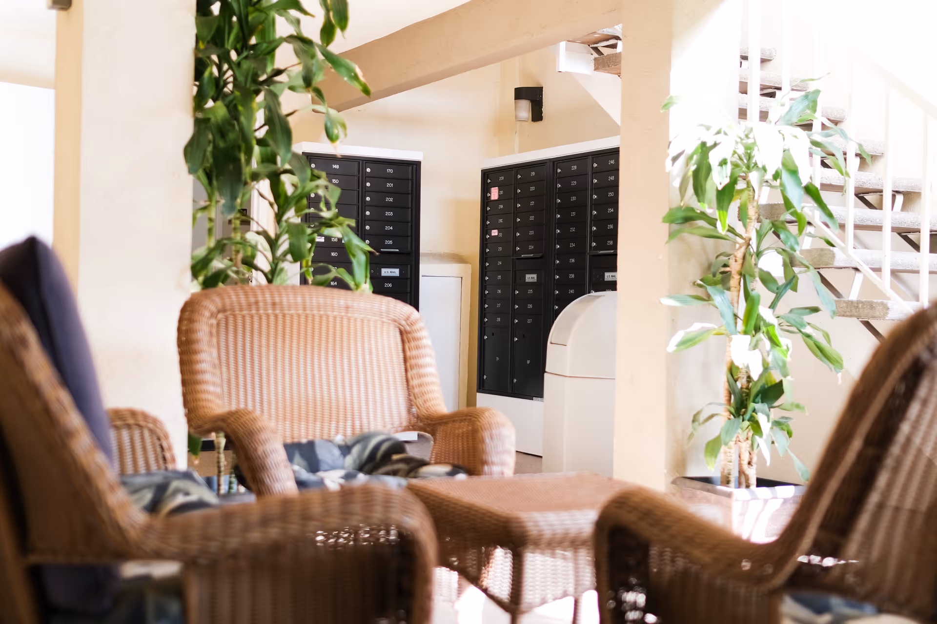 A seating area with wicker chairs and a table in a well-lit indoor space, featuring green potted plants and a wall of mailboxes in the background near a staircase.