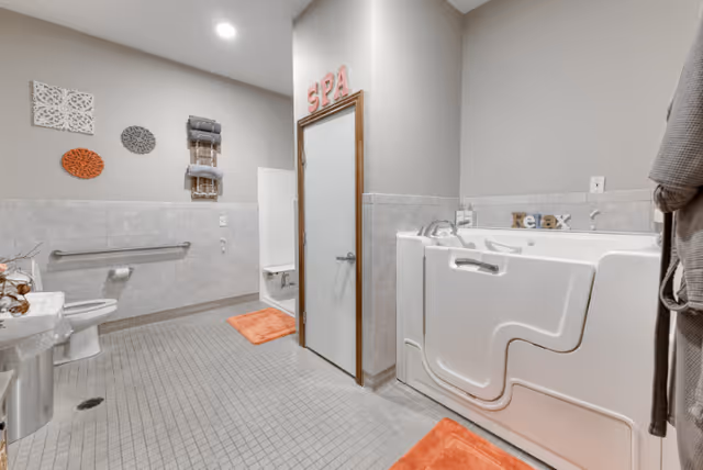 A clean, accessible bathroom with a walk-in bathtub, a toilet with grab bars, a sink, and a small shower area. The walls are tiled halfway up, and there are orange bath mats on the floor. Decorative wall hangings and towels are visible, with the word 'SPA' displayed above a door.