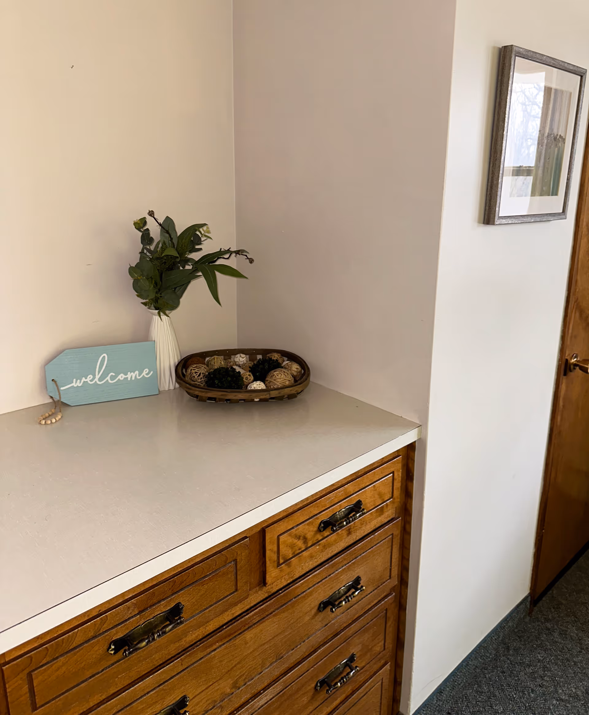 A wooden dresser with a white countertop in a corner of a room. On the countertop, there is a white vase with green leafy plants, a small blue sign that says 'welcome,' and a decorative basket filled with various natural elements. A framed picture hangs on the adjacent wall, and a wooden door is partially visible.