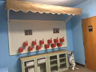 An interior room with light blue walls featuring a pegboard with red mailboxes labeled 'US MAIL' and 'POST OFFICE'. Below the pegboard is a cabinet with glass doors. To the right, there is a door with a restroom sign and a Dalmatian dog statue sitting on the floor.