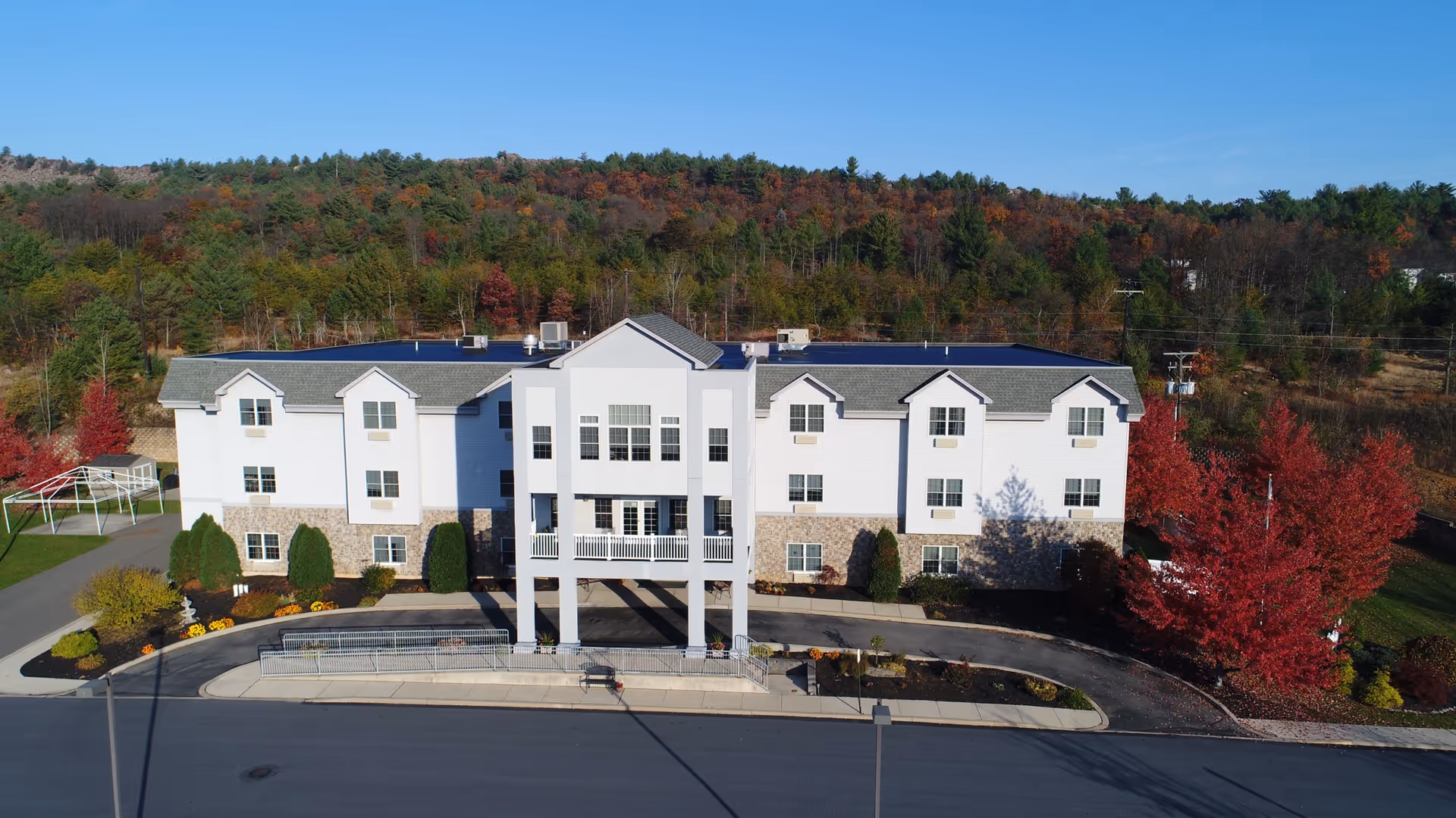 Front exterior view of a three-story senior living facility named The Palmerton, surrounded by landscaped greenery and trees with autumn foliage, set against a backdrop of forested hills under a clear blue sky.