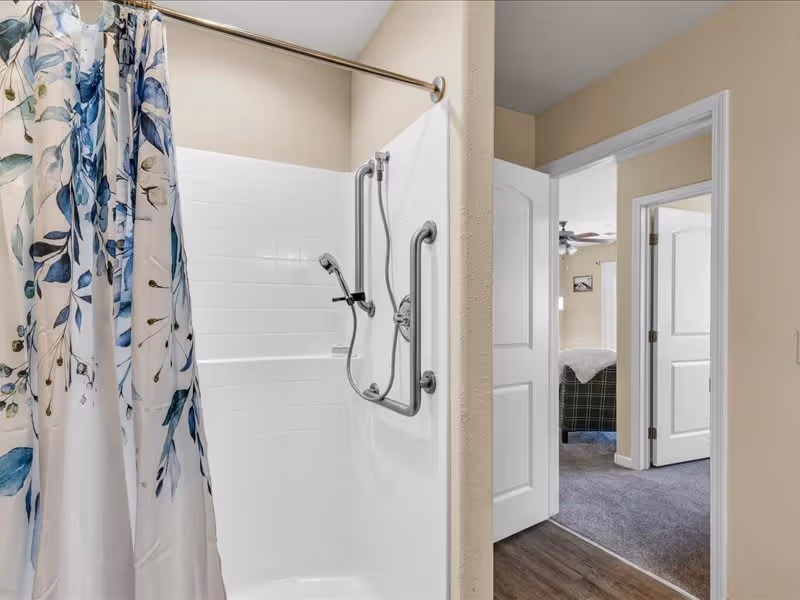 A bathroom shower area with white tiled walls and a floral shower curtain. The shower has two grab bars and a handheld showerhead. The view extends into an adjacent room with open white doors, showing carpeted flooring and part of a bed with a plaid blanket.