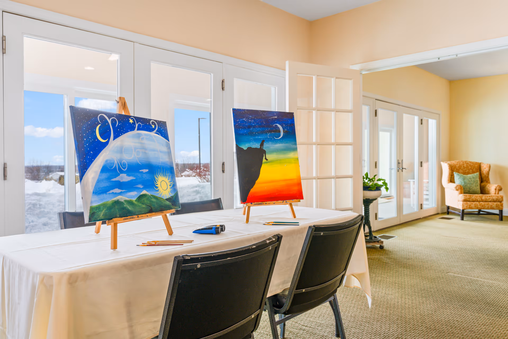 A bright room with large glass doors showing a snowy outdoor scene. Inside, a table covered with a white cloth holds two easels with colorful paintings. One painting depicts a sun over green hills with a blue sky, and the other shows a silhouette of a person sitting on a cliff against a sunset and night sky. Two black chairs are positioned at the table, and a patterned armchair with a green pillow is visible in the background.