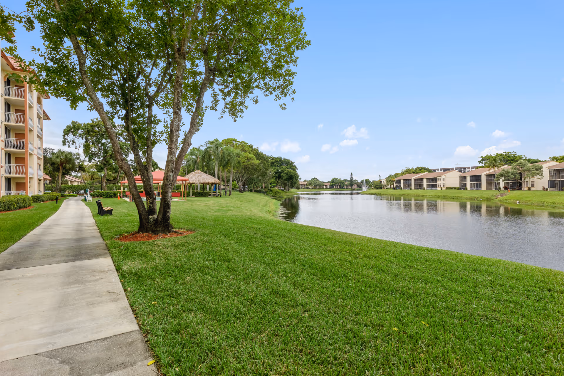 A scenic outdoor view of a senior living facility with a paved walkway alongside a grassy area and a calm pond. Trees and benches line the walkway, with residential buildings visible on both sides of the pond under a clear blue sky.