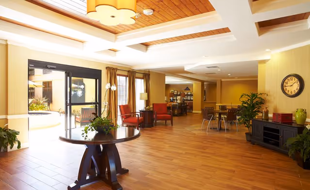 A spacious and well-lit senior living facility common area with wooden flooring, a round table with a plant centerpiece, red chairs, a clock on the wall, and large windows letting in natural light. The area includes seating and a small dining table, with a view of an outdoor patio through glass doors.