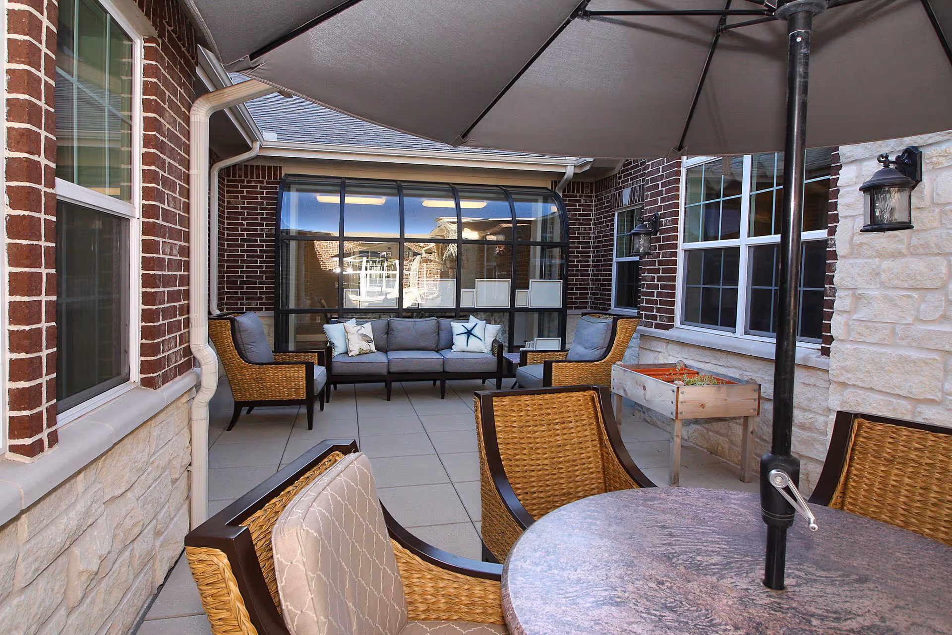 Outdoor patio area with a round table and wicker chairs under a large umbrella. In the background, there is a seating area with a gray cushioned sofa and two matching armchairs, surrounded by brick and stone walls with windows.