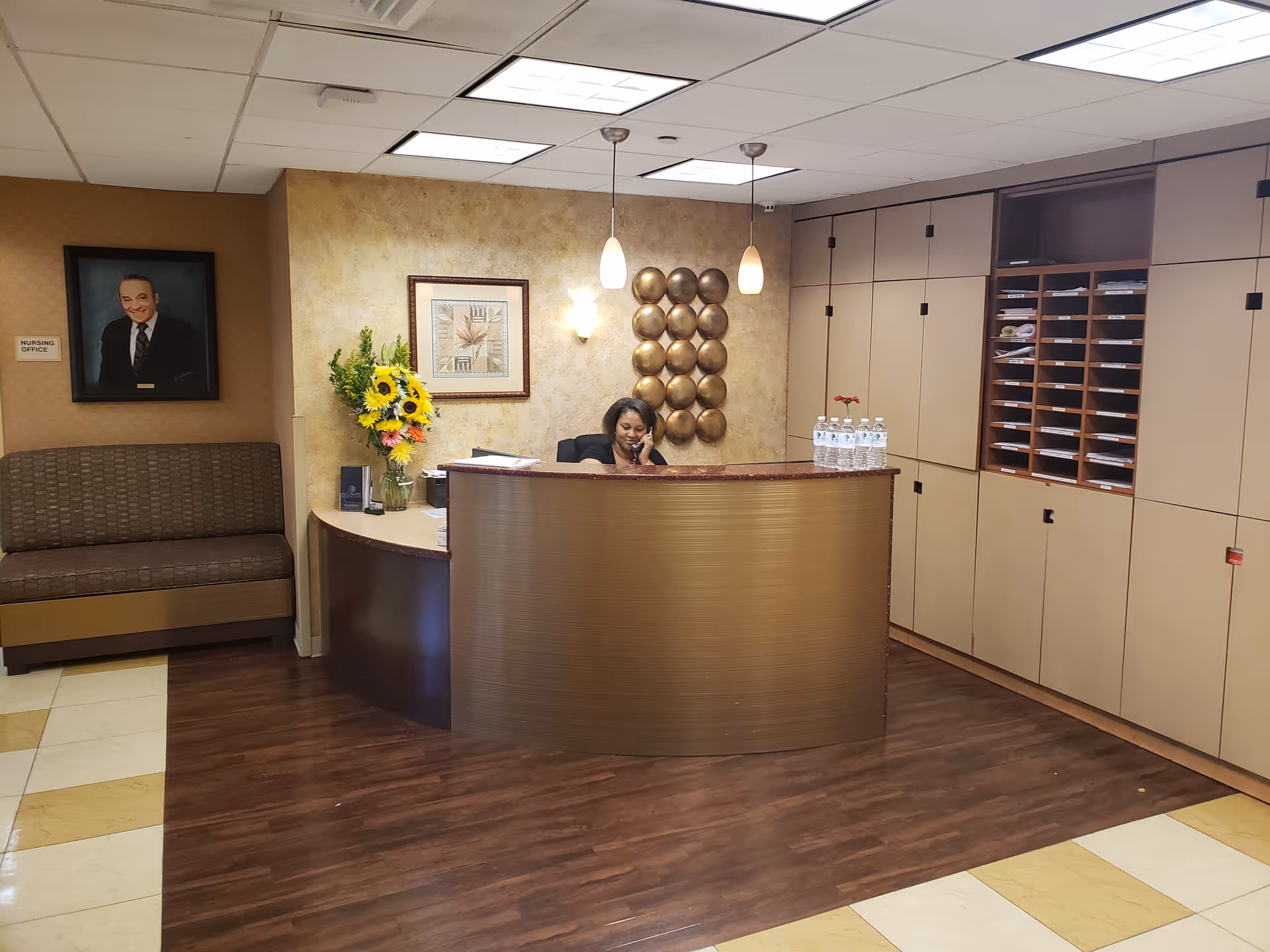 A reception desk in a healthcare facility lobby with a woman on the phone, a vase of sunflowers, seating to the left, and mail cubbies to the right.