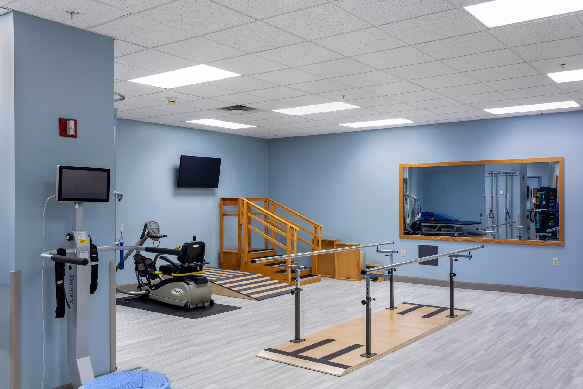 A physical therapy room with light blue walls and a light wood floor. The room contains parallel bars for walking exercises, a recumbent exercise bike, a wooden ramp and stairs for mobility training, a large wall mirror, and a mounted TV screen. The ceiling has recessed lighting panels.