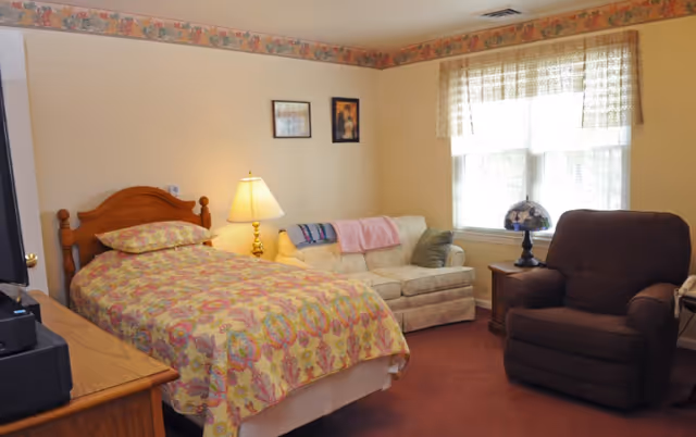 A furnished bedroom with a single bed covered in a floral bedspread, a loveseat, recliner, side table and window.