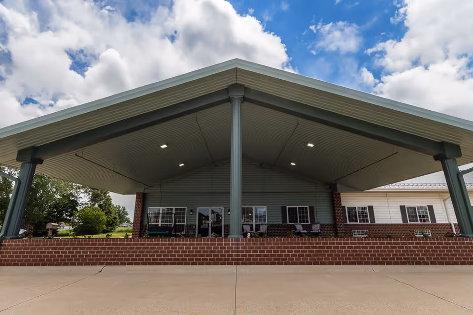 Covered outdoor patio area with a high peaked roof supported by columns, brick half-wall, and seating visible inside. The sky is partly cloudy.