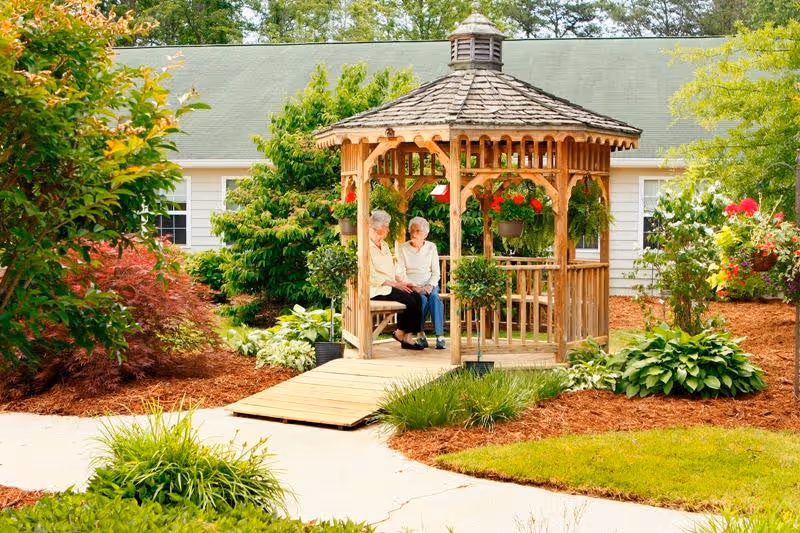 Two elderly women seated in a wooden gazebo in a landscaped courtyard at a senior living facility.