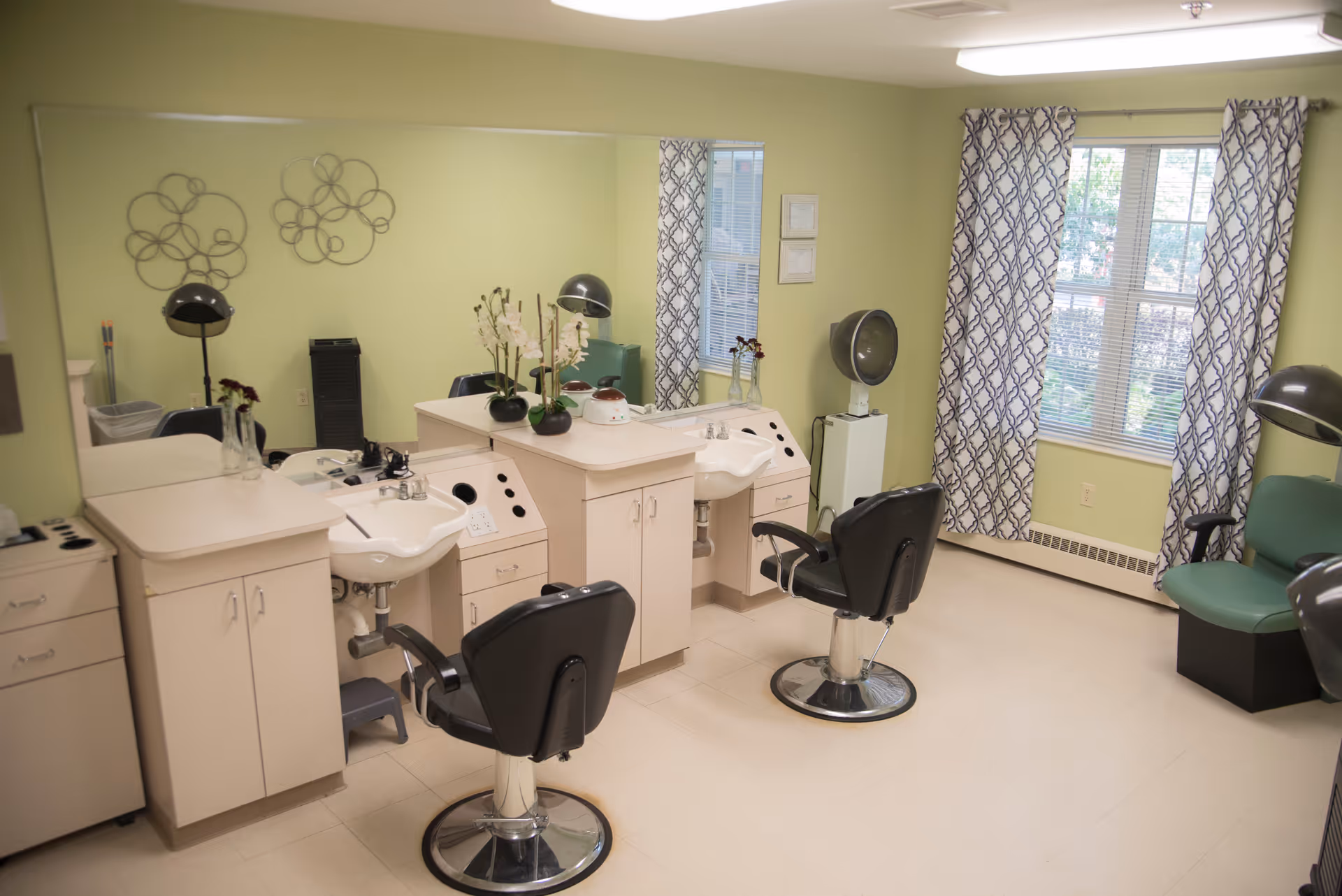 Bright salon styling room with two salon chairs, wash sinks, mirrors, hooded hair dryers, and green walls with patterned curtains.