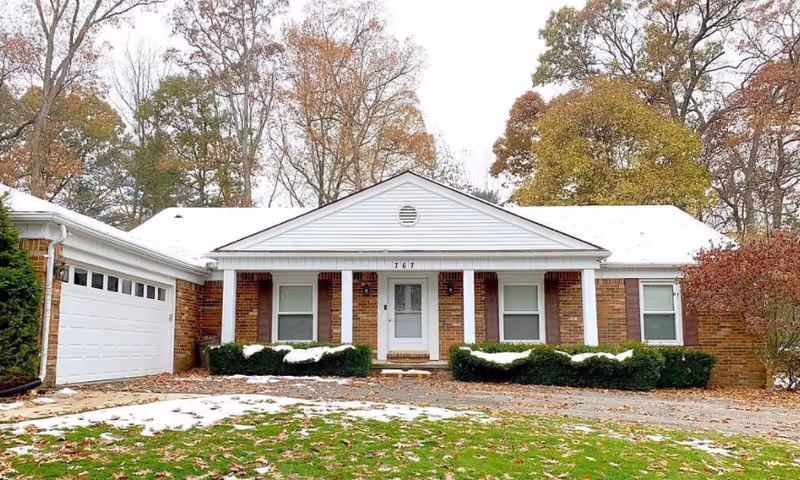 Front exterior view of a single-story brick house with a white garage door on the left and a white front door in the center. The house has a small porch with four white columns and bushes in front. There are trees with autumn foliage in the background and patches of snow on the ground.