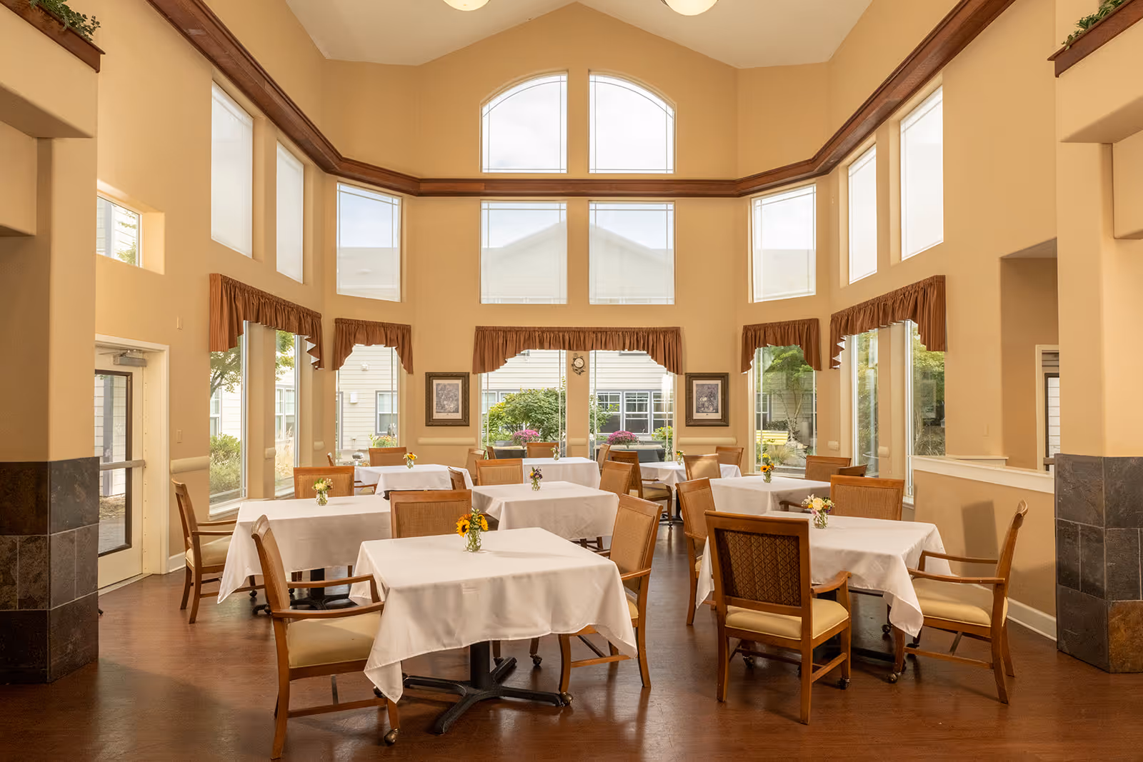 A bright dining room with multiple tables covered in white tablecloths, each adorned with small flower arrangements. The room features large windows with brown valances, high ceilings, and wooden chairs around the tables. Outside the windows, greenery and parts of the building exterior are visible.