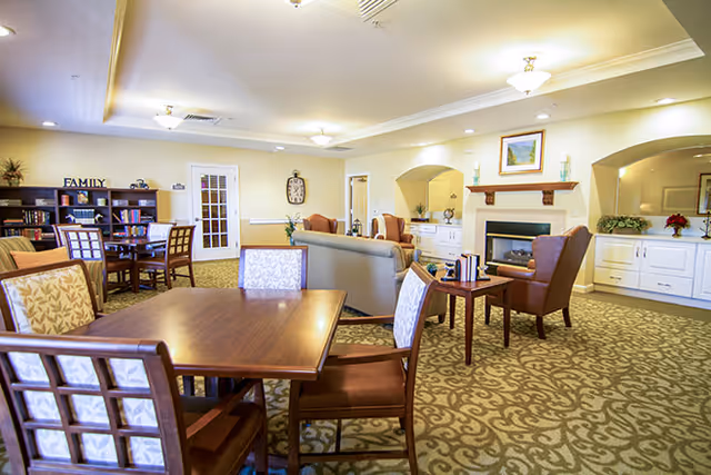 A spacious and well-lit common area in a senior living facility featuring multiple wooden tables and chairs with patterned cushions, a seating area with sofas and armchairs around a fireplace, decorative plants, bookshelves, and warm lighting fixtures on the ceiling.