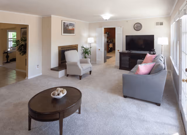 A bright living room with beige carpet, a gray sofa with pink pillows, a beige armchair, a round wooden coffee table with a decorative bowl, a flat-screen TV on a dark wooden stand, a fireplace, two floor lamps, and large windows letting in natural light.
