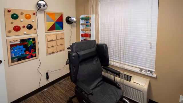 Small therapy room with a reclining wheelchair in front of a window, sensory activity boards on the wall, and an air-conditioning unit beneath the blinds.