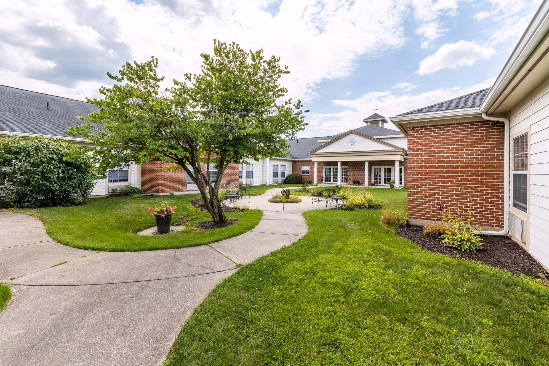 Outdoor courtyard area at Walnut Ridge Assisted Living and Memory Care featuring a curved concrete walkway, green grass, a tree in the center, potted plants, and outdoor seating with tables and chairs. The surrounding building has brick and white siding with windows and a covered entrance.