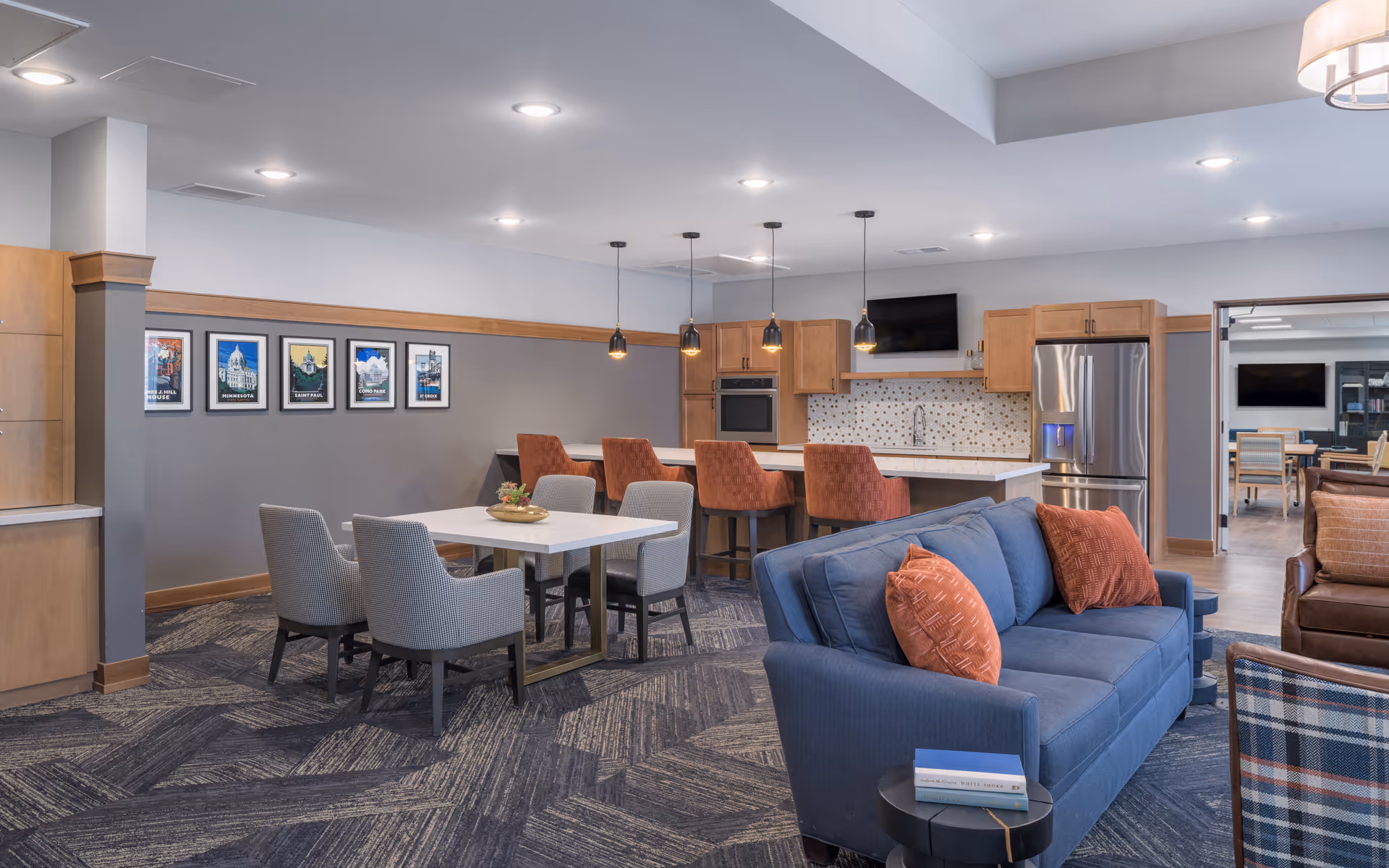 A modern senior living common area featuring a blue sofa with orange pillows, a white dining table with four gray chairs, and a kitchen area with a countertop and four orange bar stools. The kitchen has wooden cabinets, a stainless steel refrigerator, a built-in oven, and a wall-mounted TV. The room is well-lit with recessed ceiling lights and pendant lights above the counter. Framed pictures hang on the gray wall, and another seating area is visible through an open doorway.
