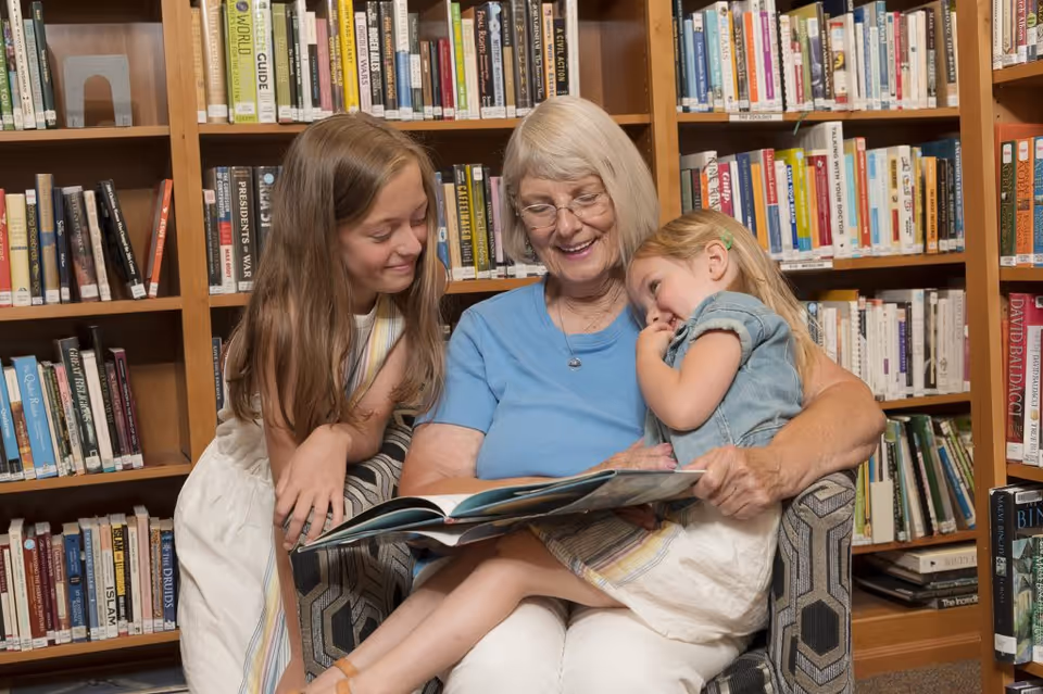 An elderly woman with glasses sitting in a chair reading a book to two young girls in a library with bookshelves filled with books in the background. One girl is sitting on her lap while the other leans in close, all smiling and engaged with the book.
