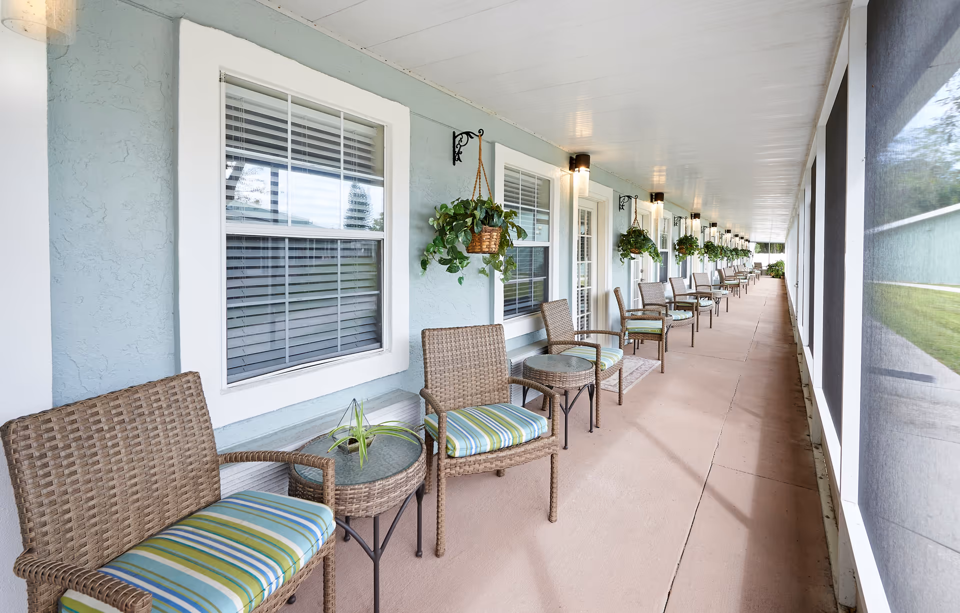Long screened porch with wicker chairs, small tables, and hanging plants along the exterior of a senior living building.