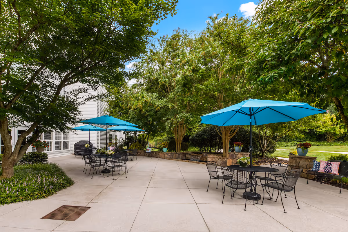 Outdoor patio area with several black metal tables and chairs, each table shaded by a bright blue umbrella. The patio is surrounded by lush green trees and plants, with a stone wall and decorative flower pots. A white building with large windows is visible on the left side.