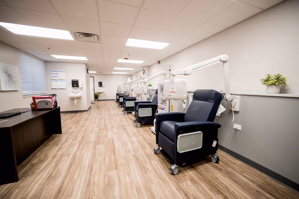 A clean, modern medical treatment room with multiple reclining chairs equipped with attached medical devices and overhead adjustable arms. The room has wood-patterned flooring, white walls with a gray lower half, bright ceiling lights, and sinks mounted on the walls. A desk with files and office supplies is visible on the left side near a window with blinds.