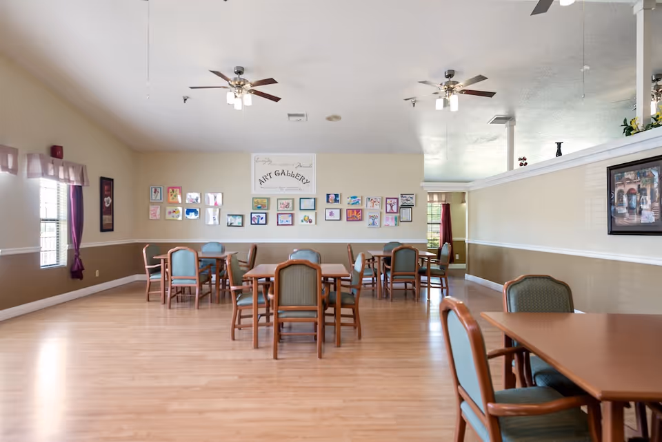 Spacious common dining/activity room with several tables and chairs, artwork on the wall and ceiling fans.