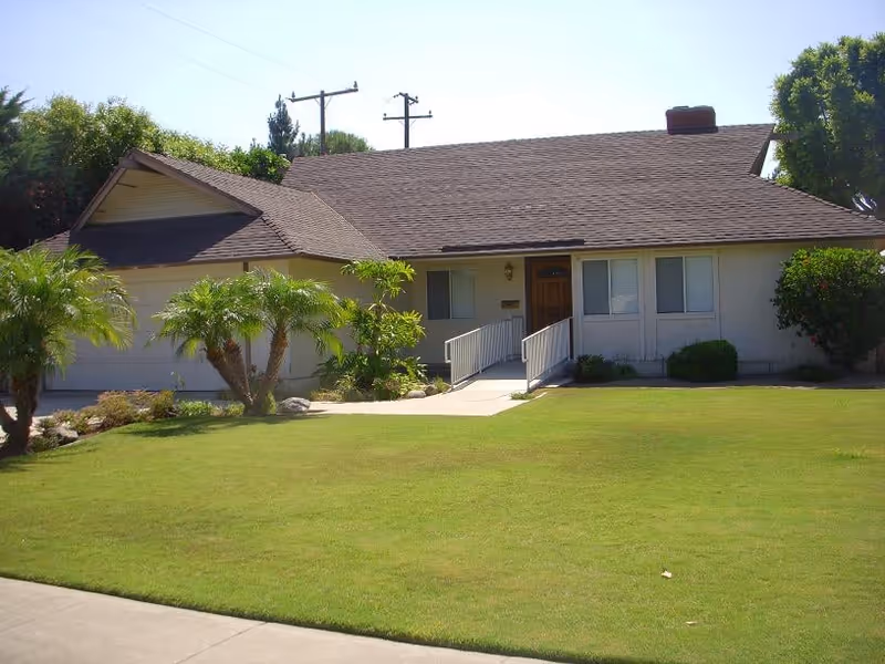 Single-story suburban house front with a wheelchair ramp, garage, and a well-kept lawn.