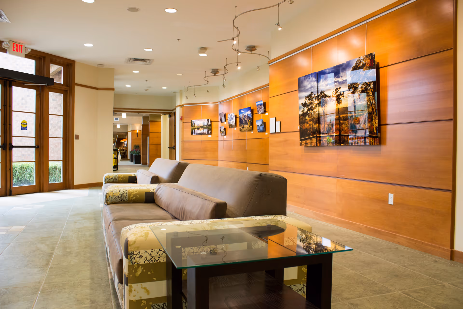 A spacious and well-lit common area with a long gray sofa and a glass-top table in front. The walls are wooden paneled and decorated with multiple framed landscape photographs. Large glass doors allow natural light to enter the space.