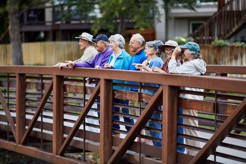 A group of seven elderly people standing on a wooden bridge outdoors, looking over the railing. One person is using binoculars, and the others are pointing or looking ahead. They are dressed in casual clothing and hats, with trees and a building visible in the background.