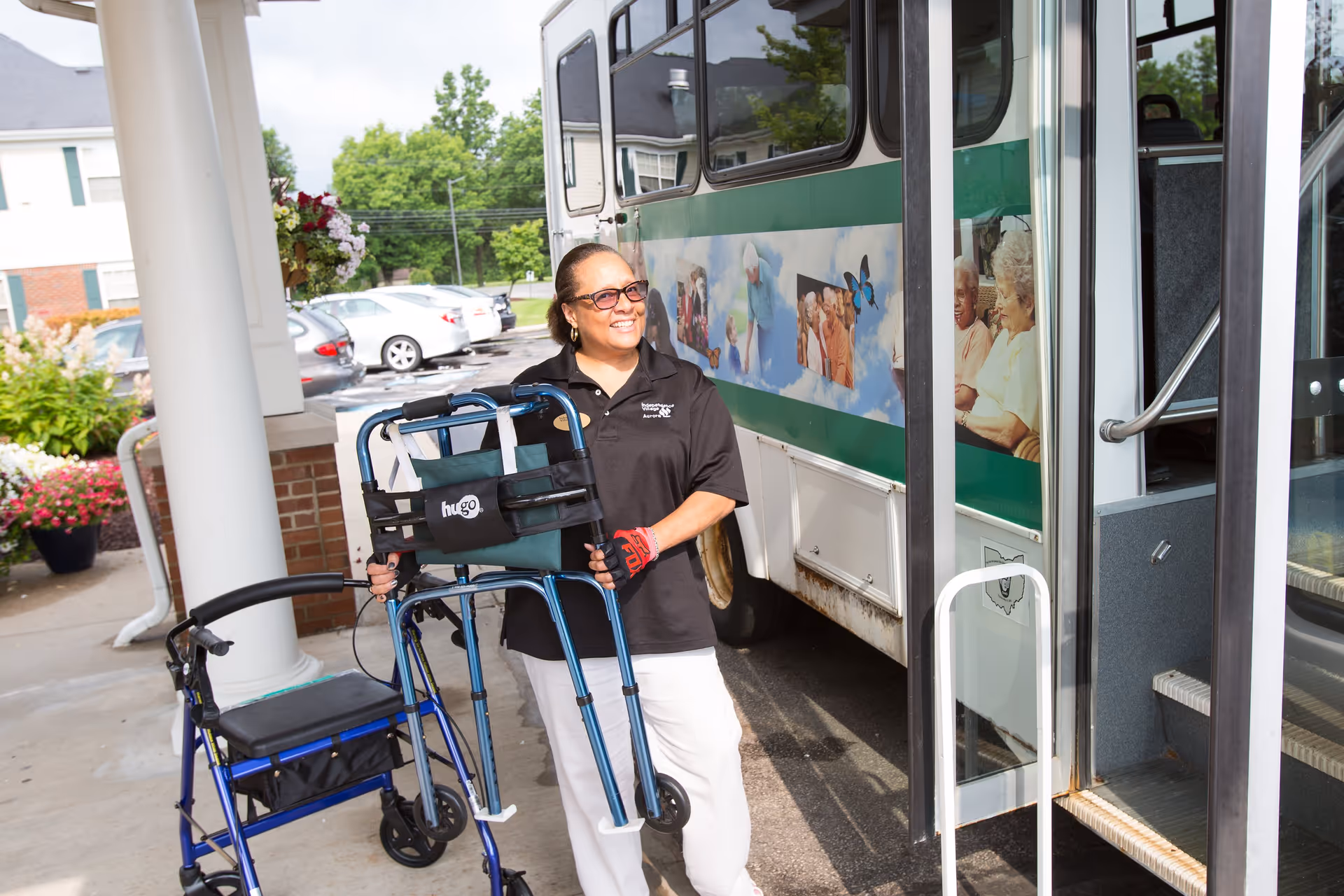 A smiling woman wearing sunglasses and a black polo shirt stands outside near a bus holding a blue walker. Another blue walker is nearby. The scene is outside a building with flowers and parked cars visible in the background.