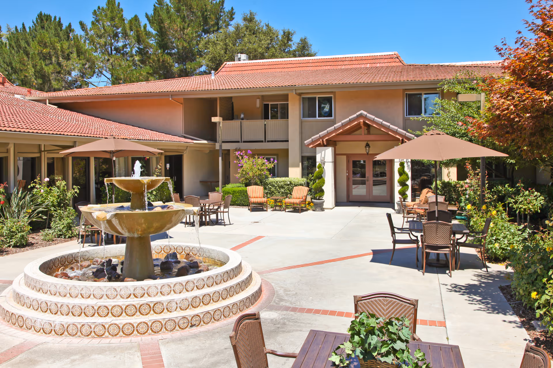 Outdoor courtyard area of a senior living facility with a multi-tiered water fountain in the center, surrounded by patio tables with umbrellas and chairs. The building has a beige exterior with a red-tiled roof, and there are trees and shrubs around the courtyard under a clear blue sky.