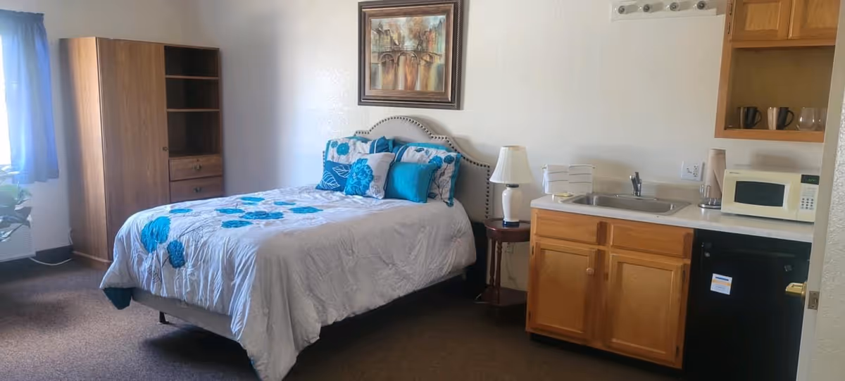 A studio bedroom featuring a bed with blue-accented bedding, a bedside table and lamp, a wardrobe, and a small kitchenette with sink, microwave and mini fridge.